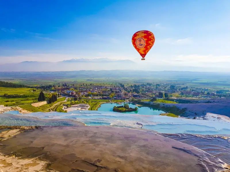 A panoramic view of Pamukkale at golden hour. There is a view of Pamukkale village, the petrified waterfall, and a hot air balloon.