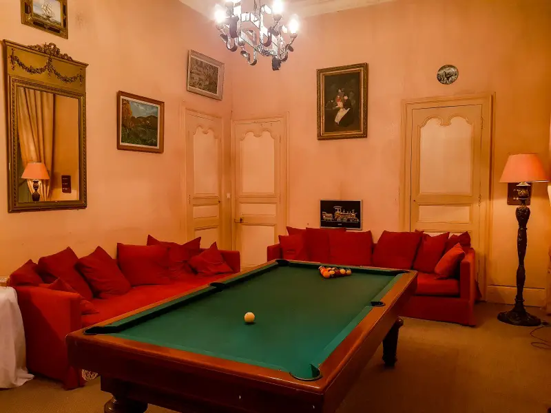 A room in a château with pink walls and a billiards table in the center. The billiards table is surrounded by red couches.