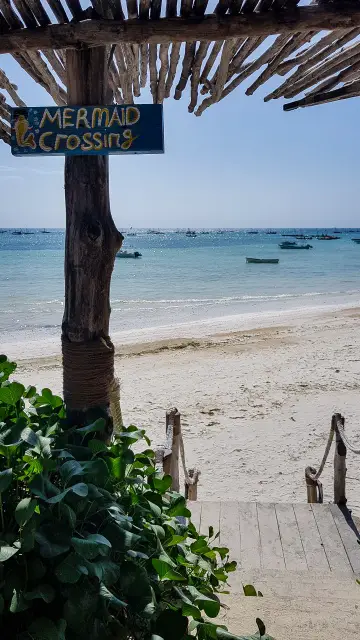 Mermaid Crossing sign hangs above a sandy beach with boats anchored in the distance - Kizimkazi, Zanzibar